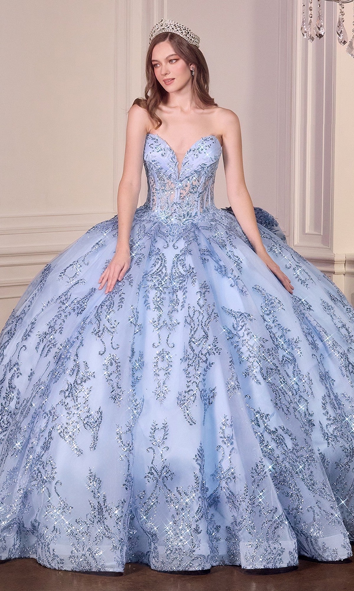 A woman stands indoors against a white wall wearing the Quinceanera Dress 15732 By Ladivine, a strapless ball gown featuring a corset bodice with lace, beaded appliqué, silver embellishments, and a sparkling tiara.